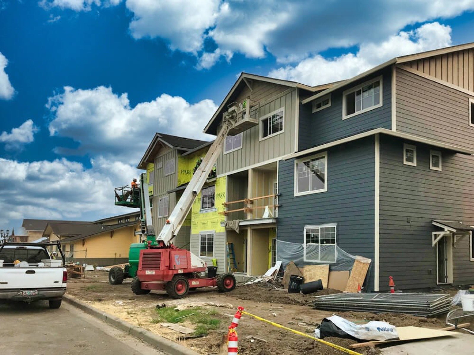 Two boom lifts beside a new three-story apartment building under construction, with blue-gray and tan siding and some walls still in house wrap