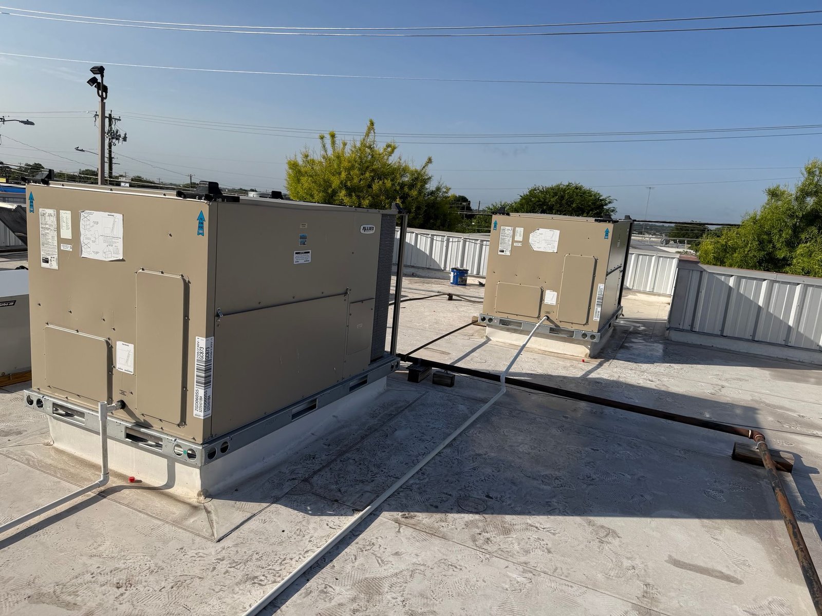 Two tan packaged rooftop HVAC units sitting on a flat commercial roof with pipes and conduit running between them