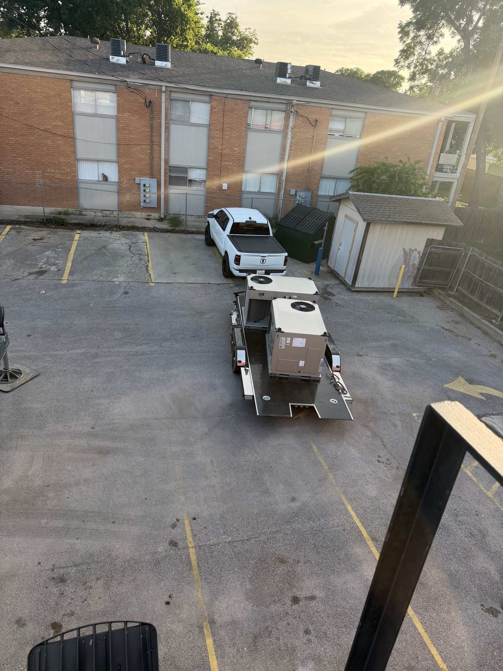 Flatbed trailer carrying packaged HVAC units parked in an apartment complex lot next to a white pickup truck