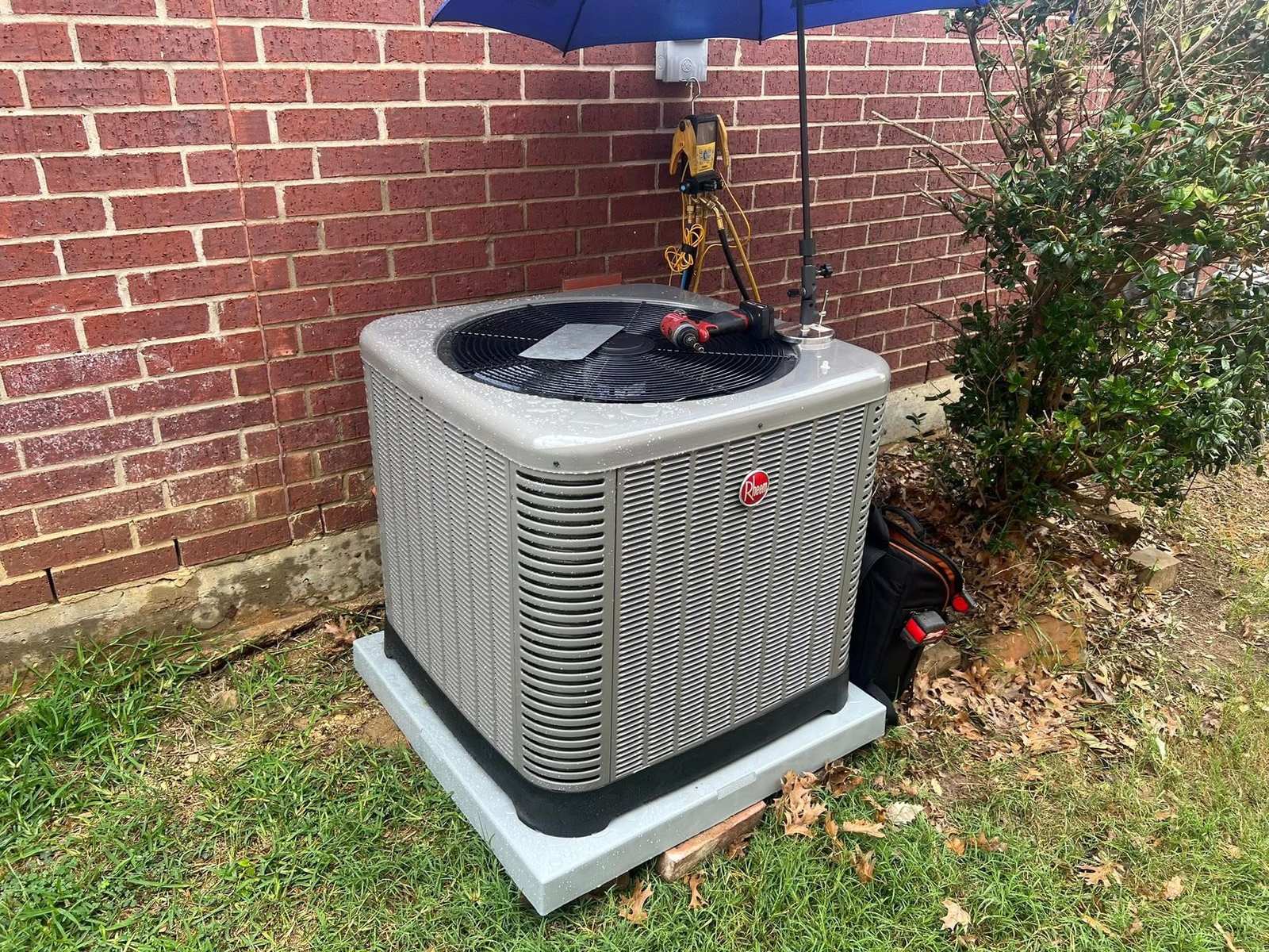 Rheem outdoor air conditioning condenser beside a brick wall, with gauges hung under a blue umbrella during service
