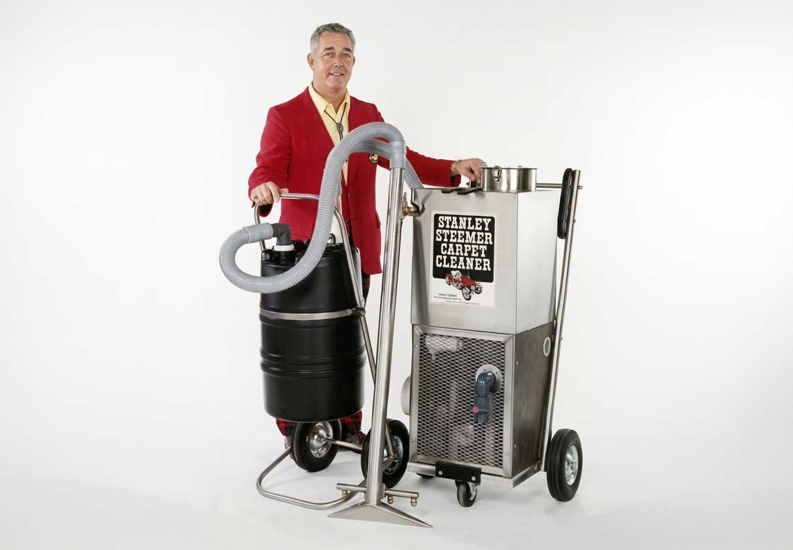 Man in a red blazer stands beside an early Stanley Steemer Carpet Cleaner, a cart-mounted machine with a hose and wand