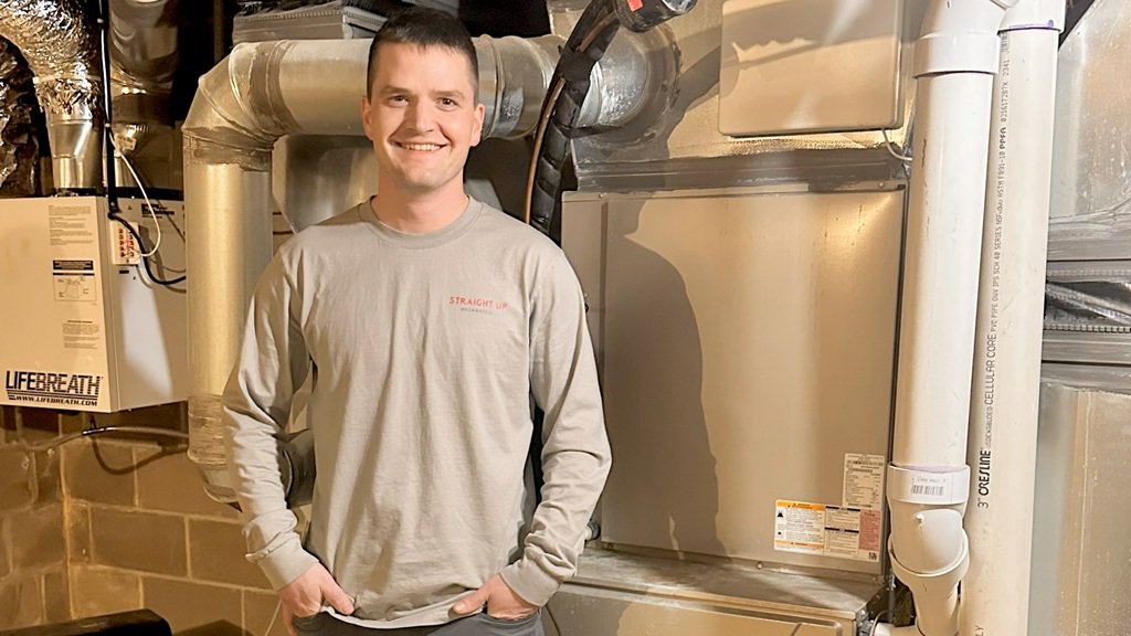 Michael Redinger, owner of Straight Up Mechanical, smiling in front of a furnace and ductwork in a basement mechanical room