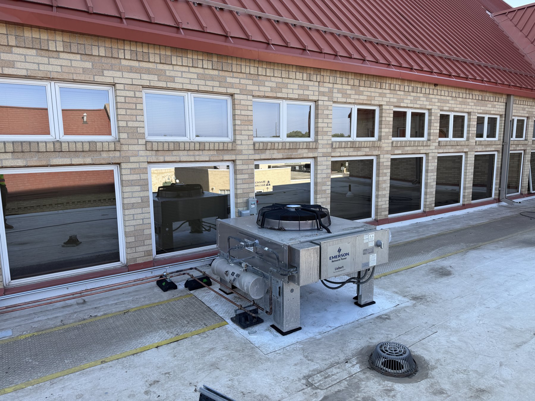 A commercial rooftop condensing unit on a flat roof beside a two-story brick building with a red metal roof