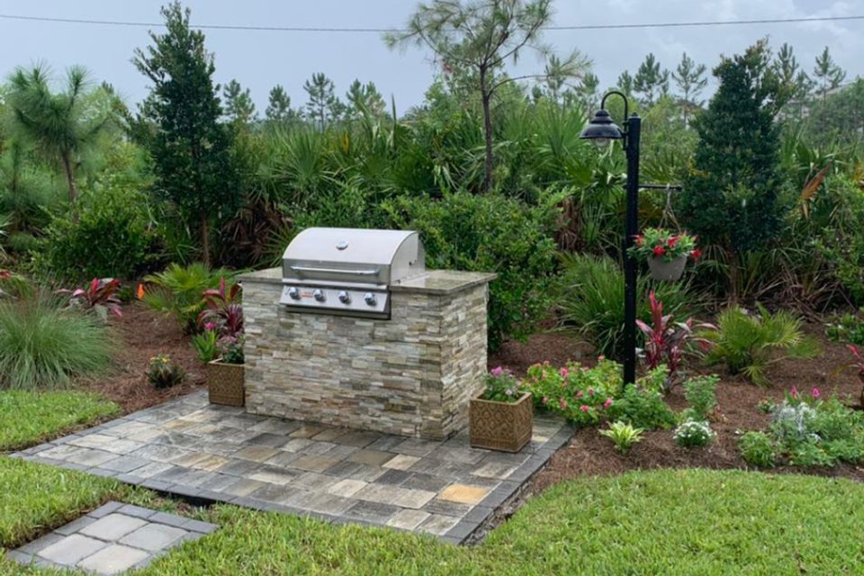 A stone grill island on a gray paver pad, surrounded by new plantings and a lamp post
