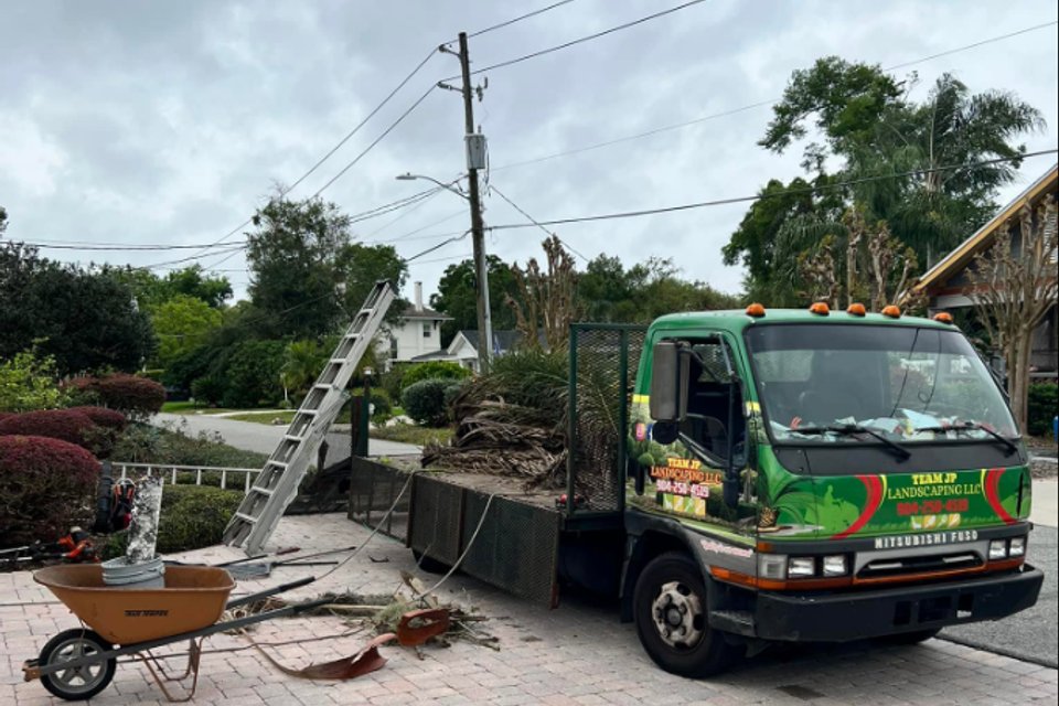 A green Team JP Landscaping truck with palm fronds in its bed, parked on a paver driveway beside a ladder and a wheelbarrow
