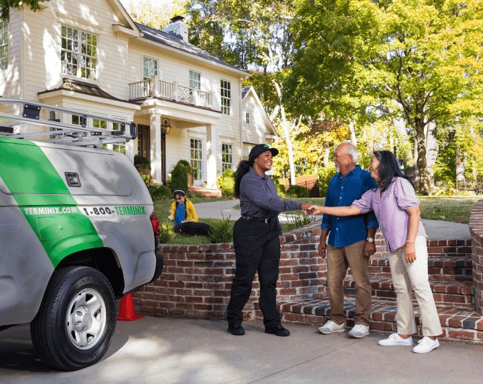 Terminix technician in uniform shakes hands with an older couple beside a brick wall outside a house