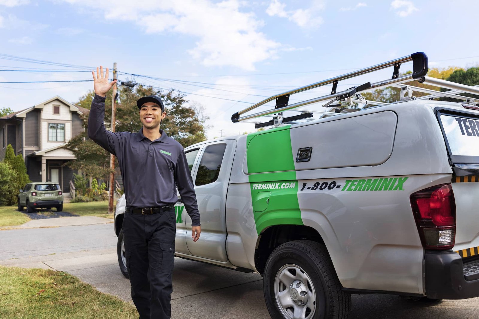 Smiling Terminix technician in dark uniform and cap waves beside a gray company pickup with a ladder rack and green logo
