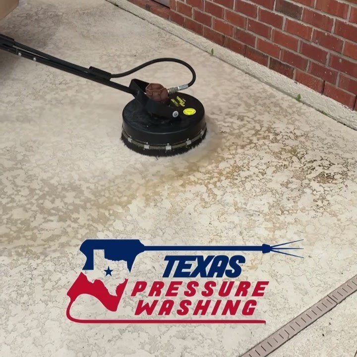 Surface cleaner with a round black housing scrubbing a stained concrete patio beside a brick wall