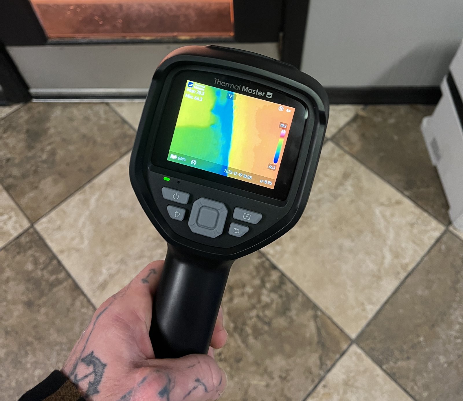 A tattooed hand holding a Thermal Master camera over a tile floor, its screen showing a blue streak across a yellow and orange image