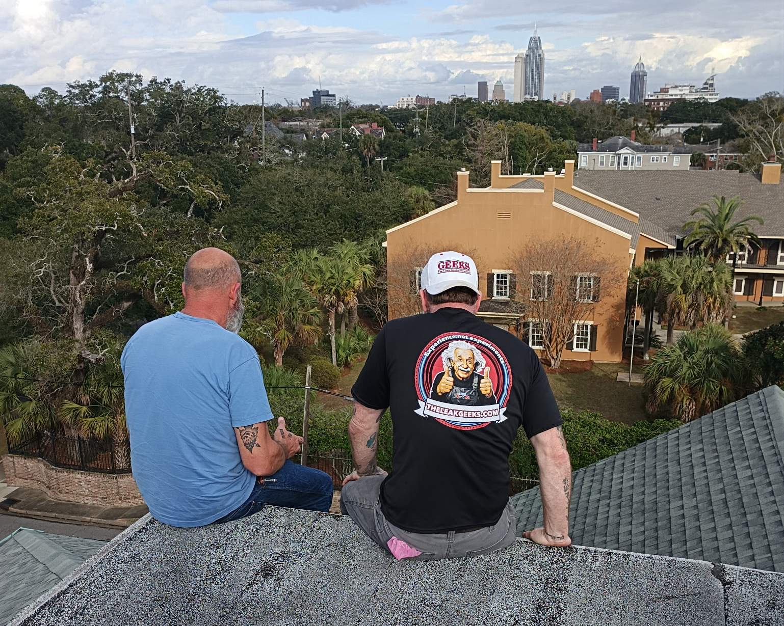 Two men sitting on the edge of a flat roof, one in a The Leak Geeks shirt and cap, with the downtown Mobile skyline in the distance