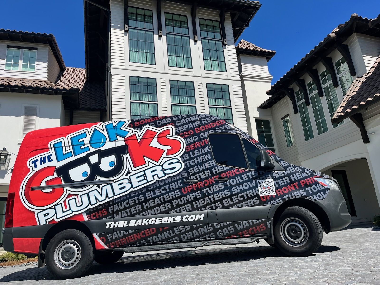 A van wrapped in red, black and white The Leak Geeks Plumbers graphics, with rows of plumbing services printed across its side, parked in front of a large white house