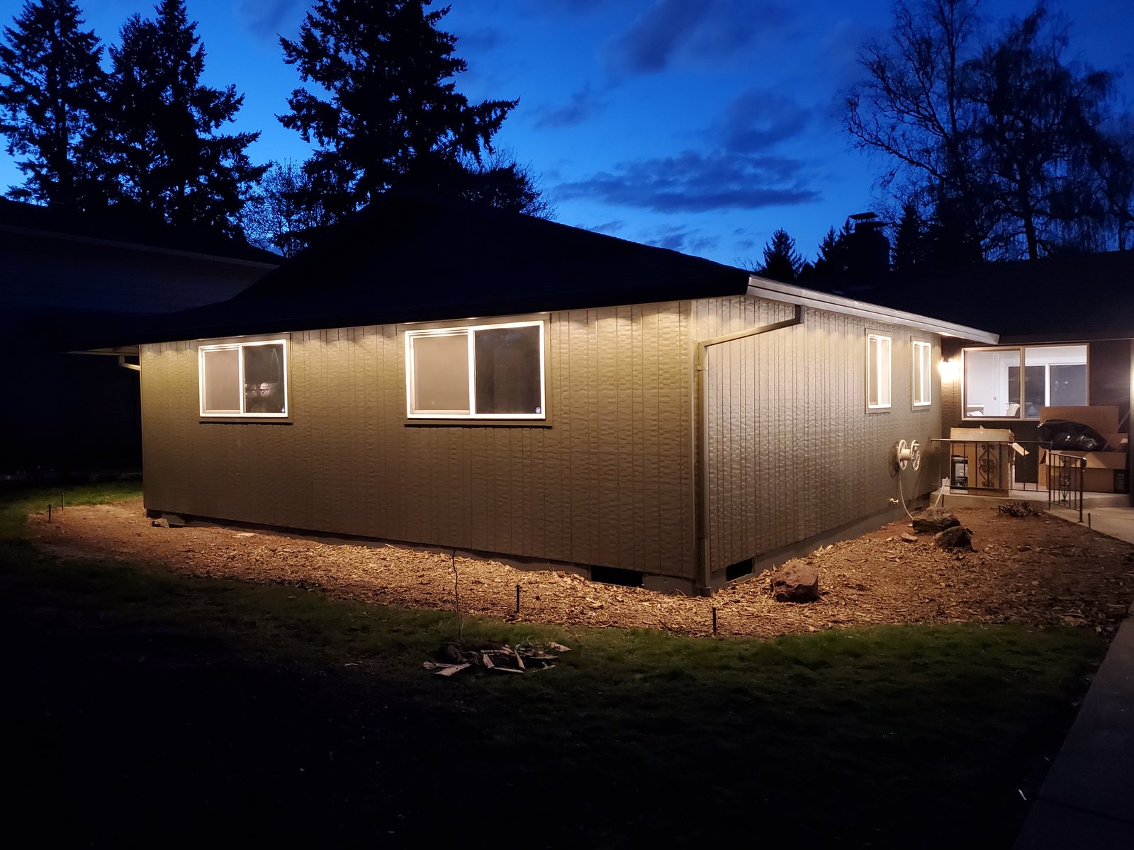 A single-story house at dusk with lights under its eaves shining on the siding, tall firs behind