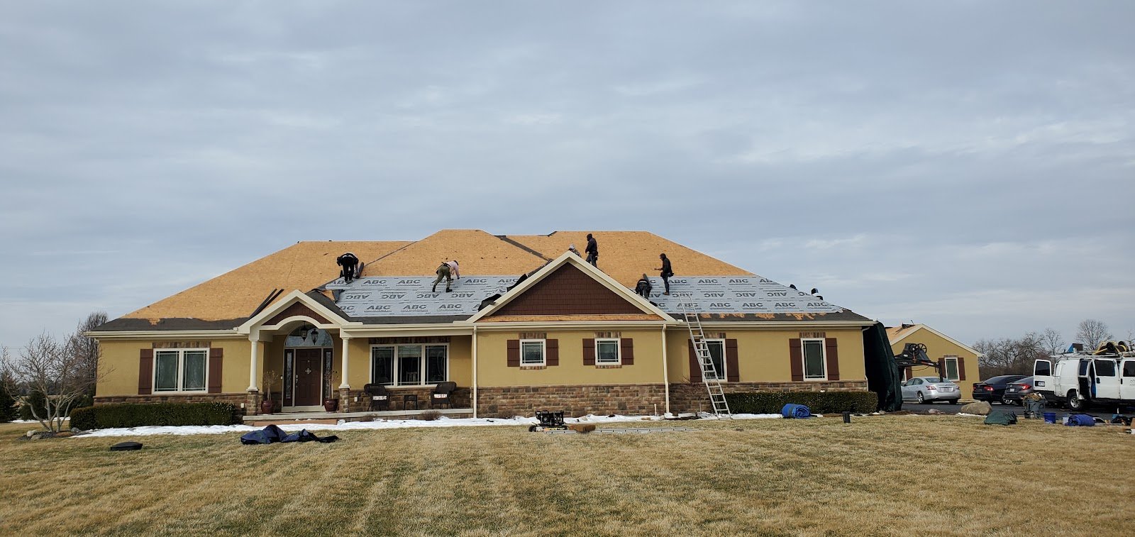 Crew members work on a partially tarped roof of a large single-story brick and stucco home, with a ladder and work van nearby