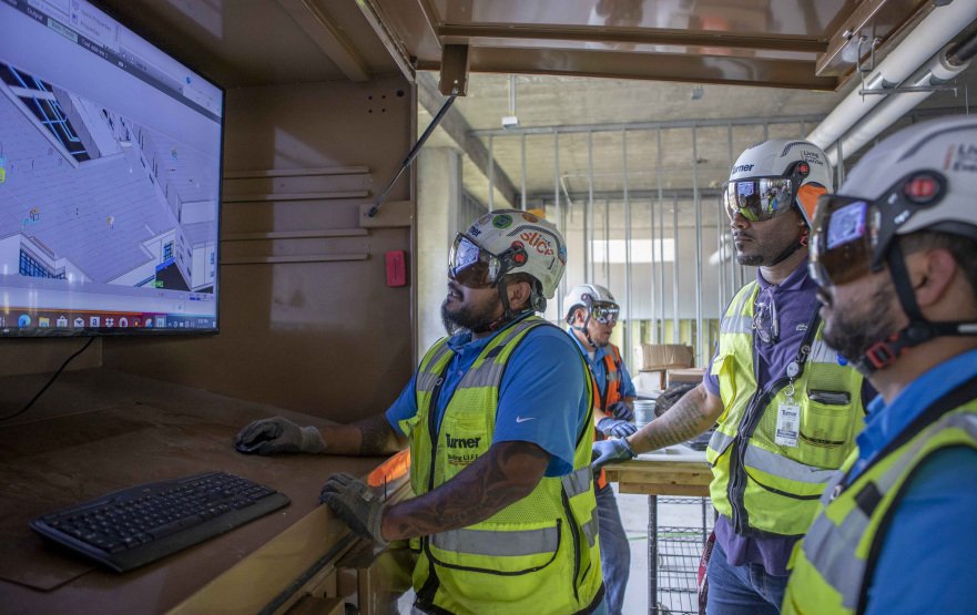 Turner workers in hard hats and safety glasses study a building model on a wall-mounted screen inside an unfinished building