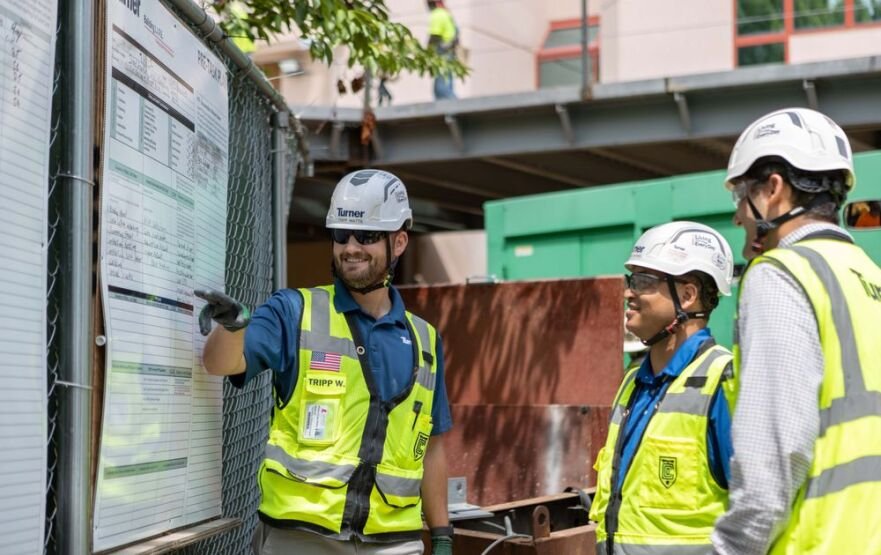 Three Turner crew members in hard hats and safety vests review a posted schedule board at a construction site