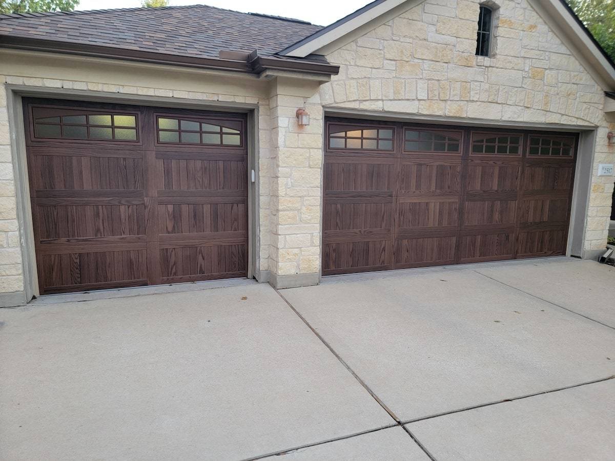 Two brown wood-look garage doors with arched windows on a stone-front house with a wide concrete driveway