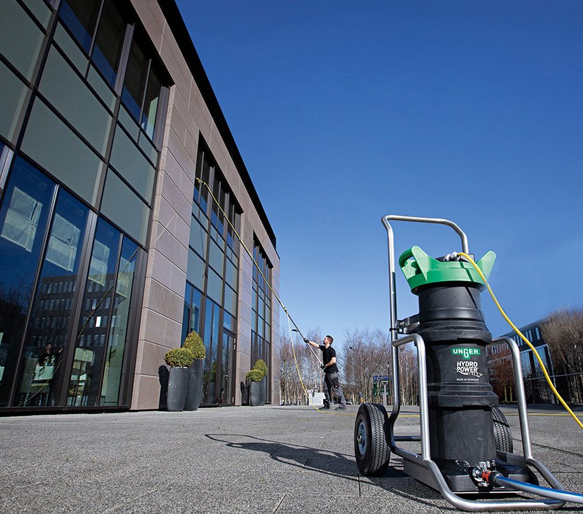 Window cleaner works a long water-fed pole against a glass office building beside an Unger HydroPower filter cart