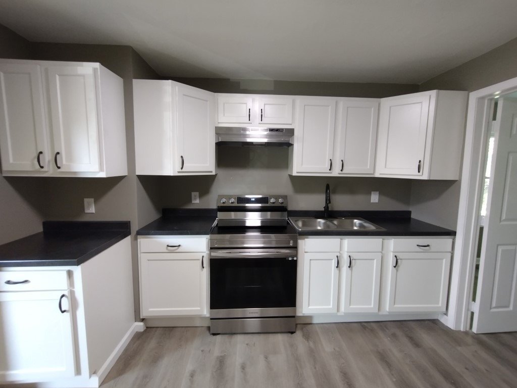 An empty kitchen with white cabinets, dark countertops, a stainless steel range and gray plank flooring, from the company's job photos