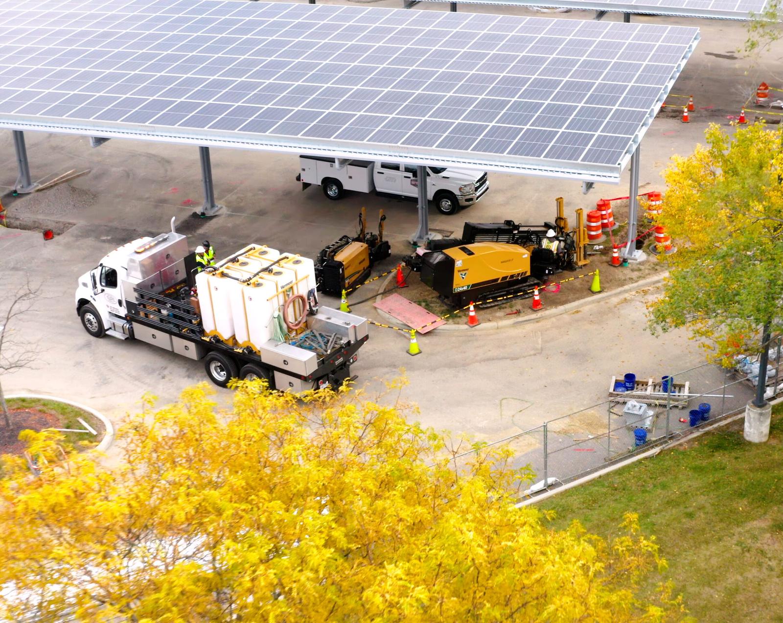 Seen from above, a yellow Vermeer D24x40 horizontal directional drill and a flatbed truck carrying white water tanks, set up beside a solar carport with orange cones and barrels around the bore site