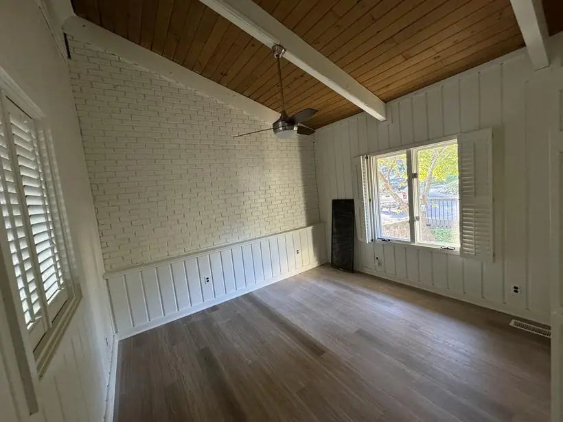 Room with white-painted brick wall, white vertical paneling, exposed beams and wood ceiling, ceiling fan and shuttered window