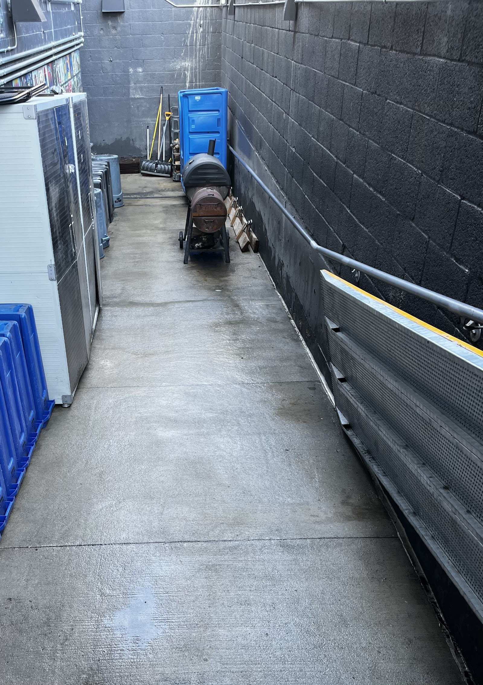 Damp concrete ramp between two walls, with brooms, a blue portable unit and other equipment at the far end