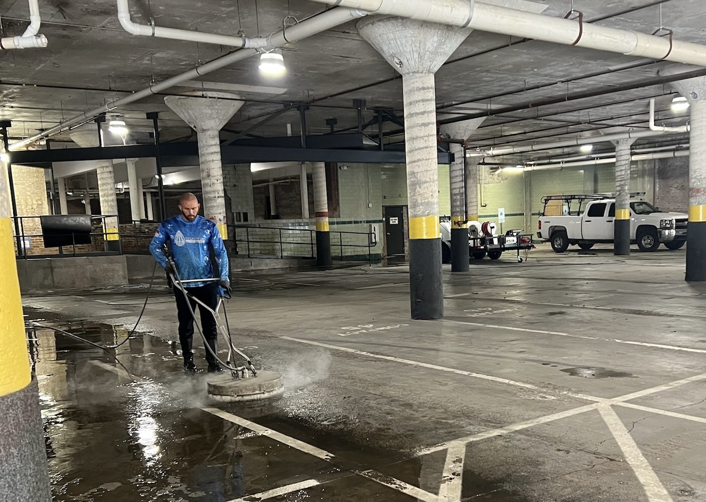 Worker in a blue jacket running a surface cleaner across the wet concrete floor of a parking garage