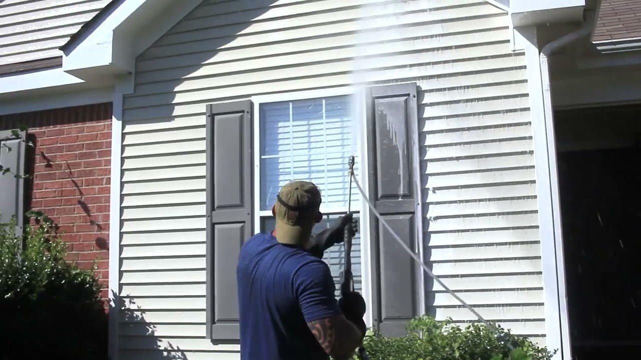 Man in a cap and blue T-shirt spraying the vinyl siding and shutters of a house with a wand
