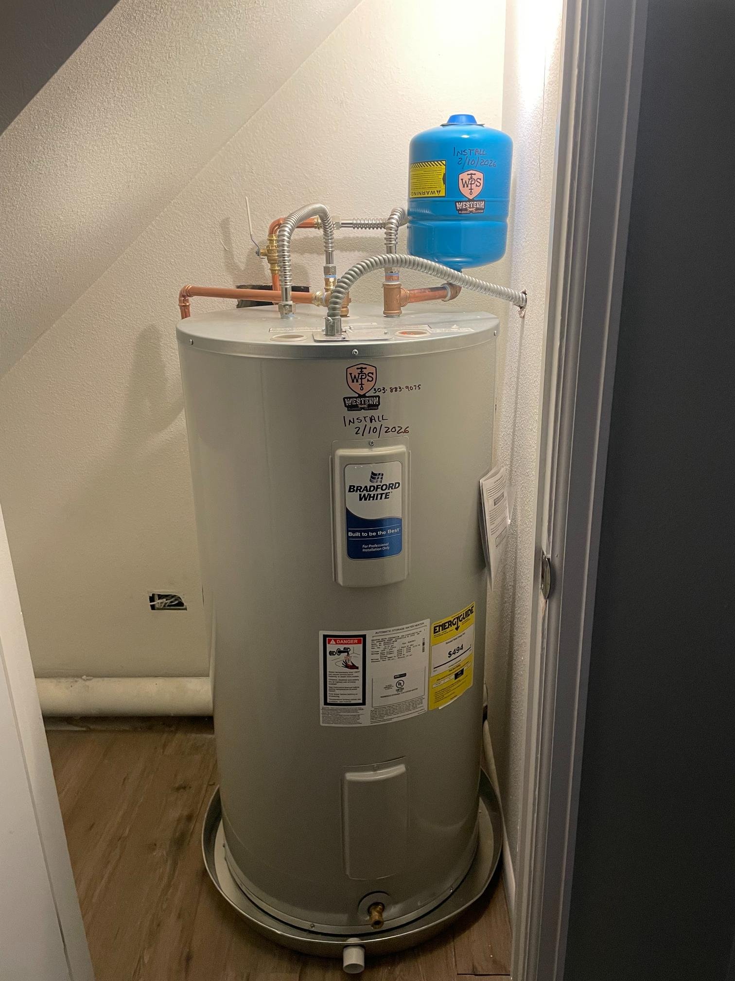 Electric water heater in a closet with copper and flexible connections, an expansion tank on top, and an install date written on the front