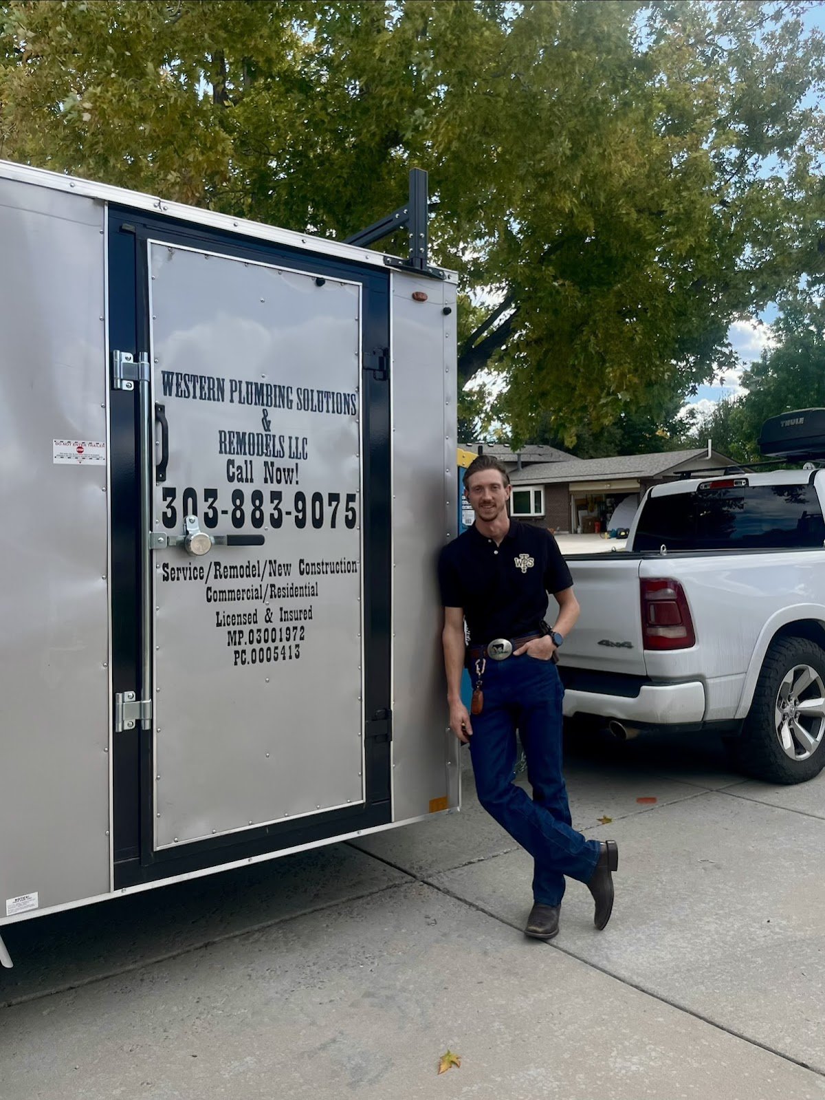 Man in black shirt and jeans leans against a silver work trailer lettered Western Plumbing Solutions & Remodels, beside a white pickup