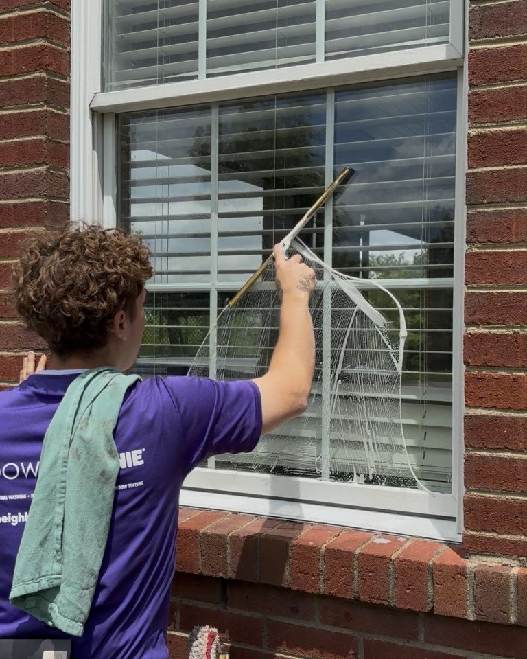 Window cleaner in a purple company shirt squeegees a soapy window set in a red brick wall